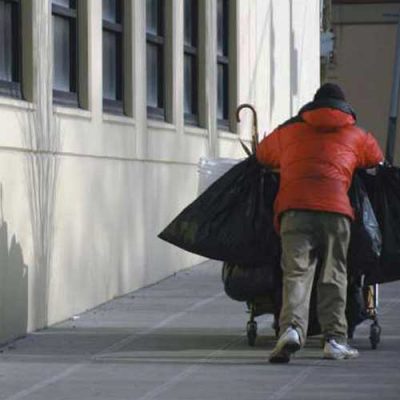 Back of a homeless man, wearing an orange jacket, pushing a cart on the sidewalk next to a building.